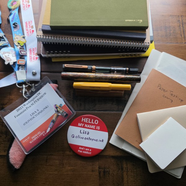 A stack of A5 and B6 notebooks, pen show lanyards and name badge, three fountain pens on a wooden pen stand, stack of hand-made booklets in multiple sizes and shapes, and a red and white 'I'm a Pen Addict' button for Lisa @oliveoctopus.ink.