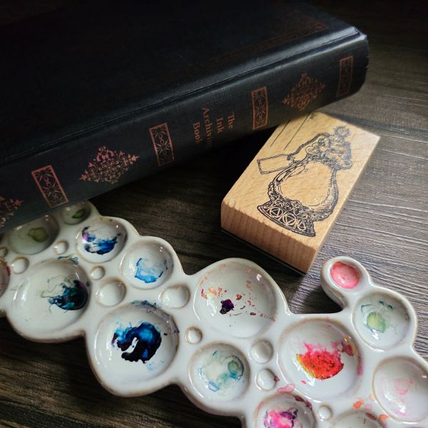 A B6 hardcover book with black cover and foil decoration and the text 'The Ink Archiving Book' on the spine, next to a ceramic ink palette that looks like a cluster of bubbles of different sizes, with colorful dried ink in the wells, and a wooden rubber stamp in the shape of an intricate bottle.