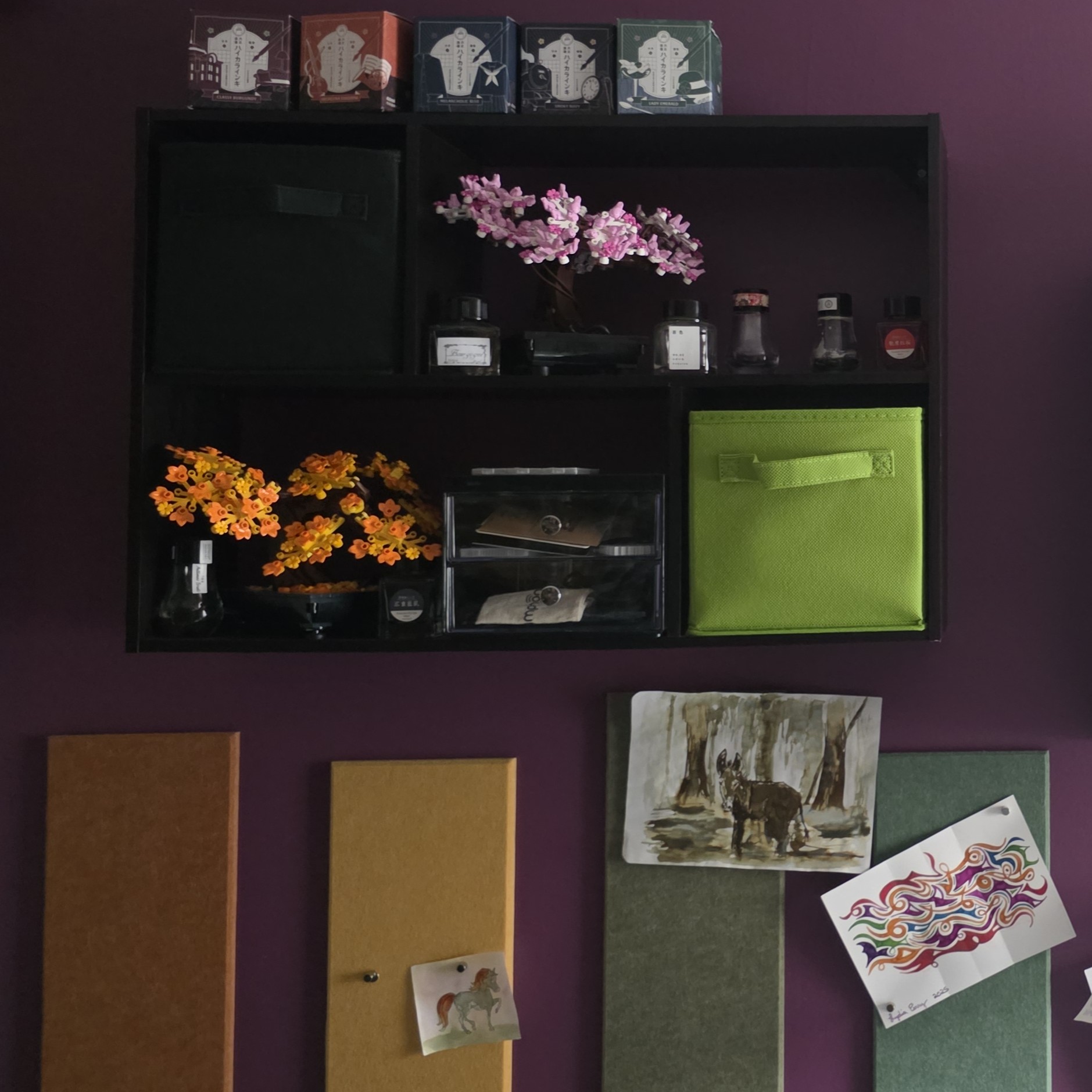 Dark brown wall shelf with four compartments arranged in a wide rectangle holding Lego bonsai trees, multiple ink boxes and empty ink bottles, and storage drawers. Below the shelf are colorful felt panels attached to the wall displaying papers with art.