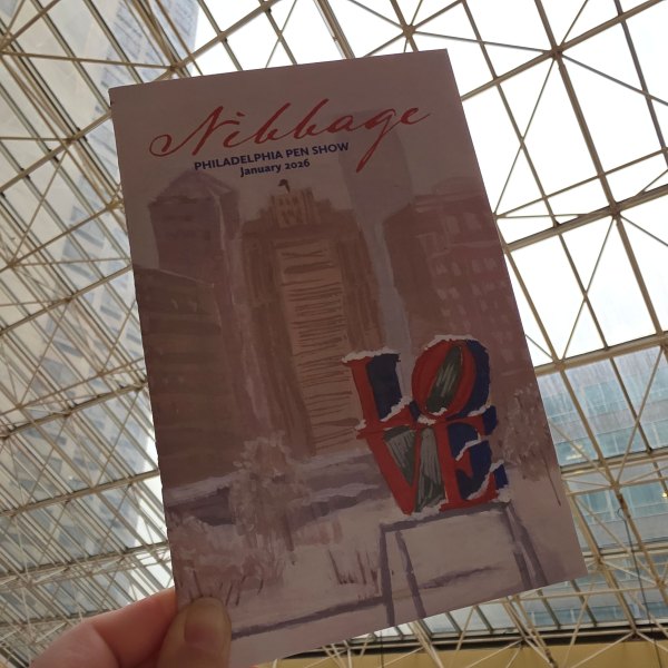 The cover of the 2026 Philly Pen Show booklet Nibbage held in front of the skylights of the show hotel.