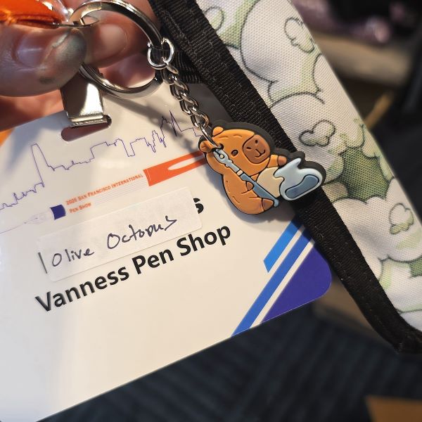 Badge for Olive Octopus, Vanness Pen Shop on a San Francisco Pen Show lanyard with a pen sleeve and small charm of a capybara with a pen and ink pot.