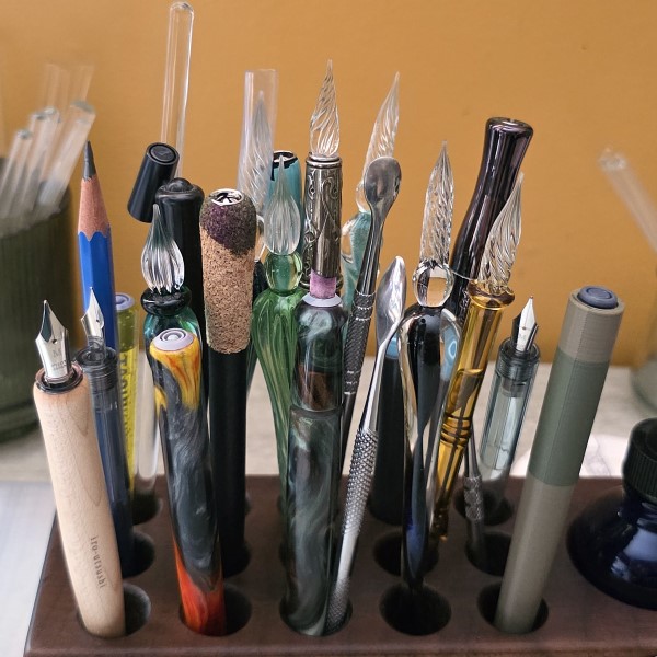 A wooden block with holes to store writing tools filled with glass pens, nib holders, metal dip pens, a long glass stir rod, metal carving spoons, and a pencil.