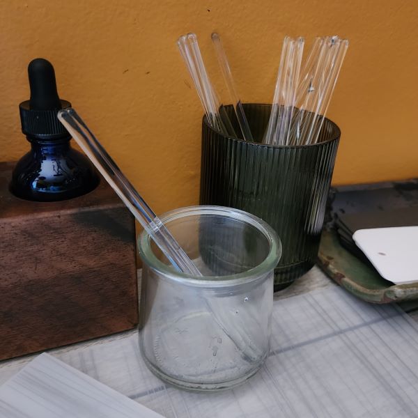 Items on a desk including a cup of glass stir rods, a jar of stir rods in water, a wood block with a blue dropper bottle, a ceramic tray with swatch cards, and a desk mat.