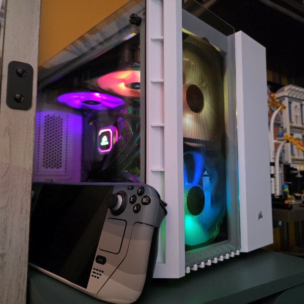 A square, white PC enclosure with windows on the front and side with colorful RGB fans and cpu cooling unit, and a Steam Deck portable game console.