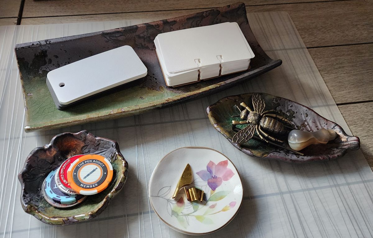 A wide rectangular ceramic piece with dark brown and green glaze holding blank Col-o-dex and Col-o-ring cards, two smaller matching pieces in the shape of different types of leaves holding challenge coins and poker chips, a bee-shaped page holder, a stone handle for wax seals, and a small painted white dish holding a folded nib and a piece that turns a straight dip pen holder into an oblique holder.