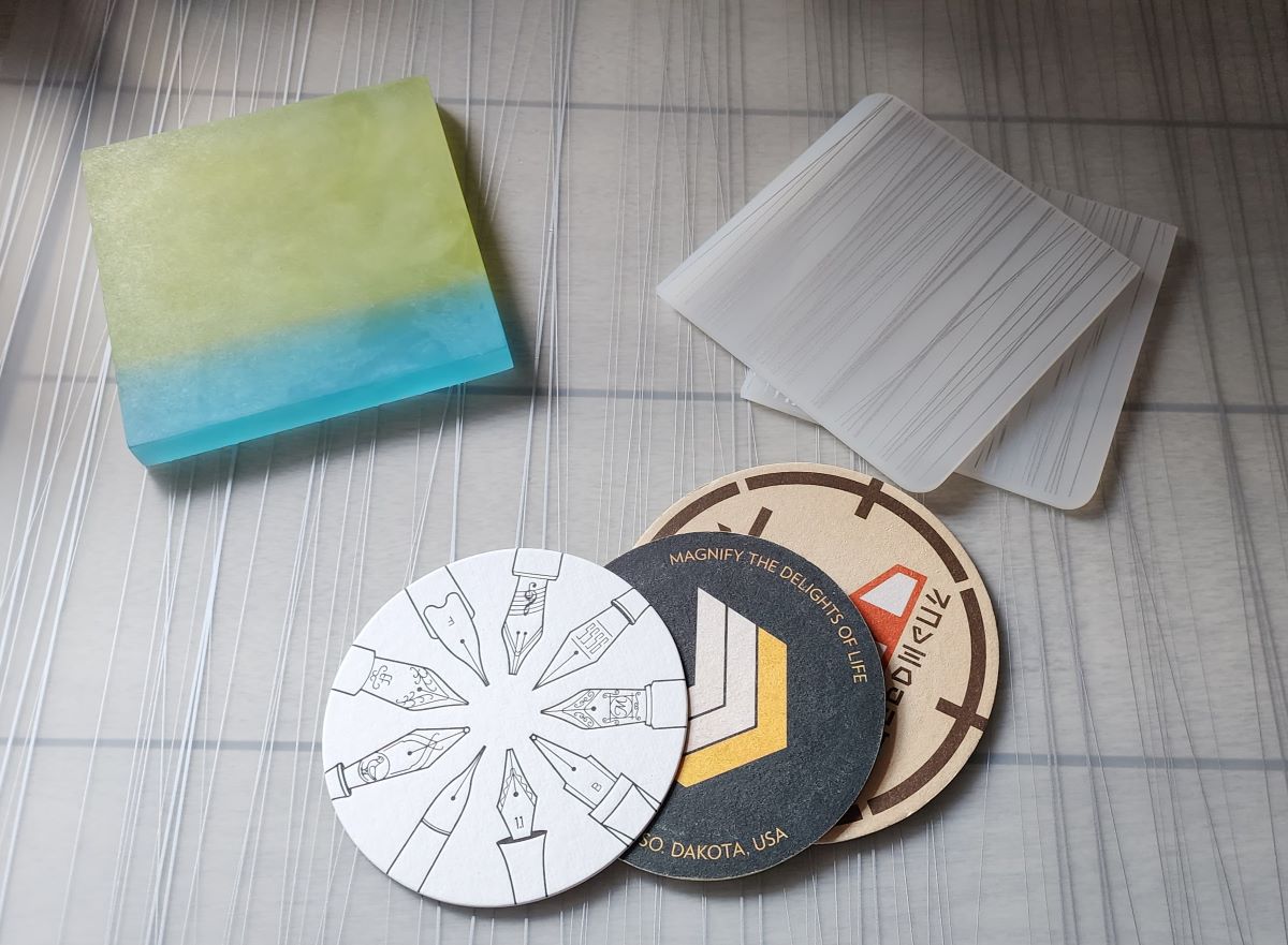 A stack of round paper coasters, the top coaster is a letterpress design with outlines of different pen nibs around the edge, thicker square silicone coasters in translucent white with a pattern of fine, silver vertical lines, and a thick acrylic coaster in frosted blue and green colors.