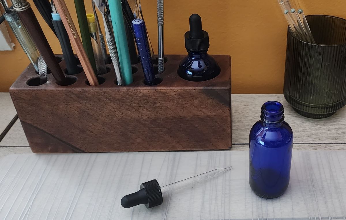 Two small cobalt blue glass bottles with dropper caps, one uncapped and the other perfectly fit into a slot of a wooden block that also holds various dip pens and swatching tools.