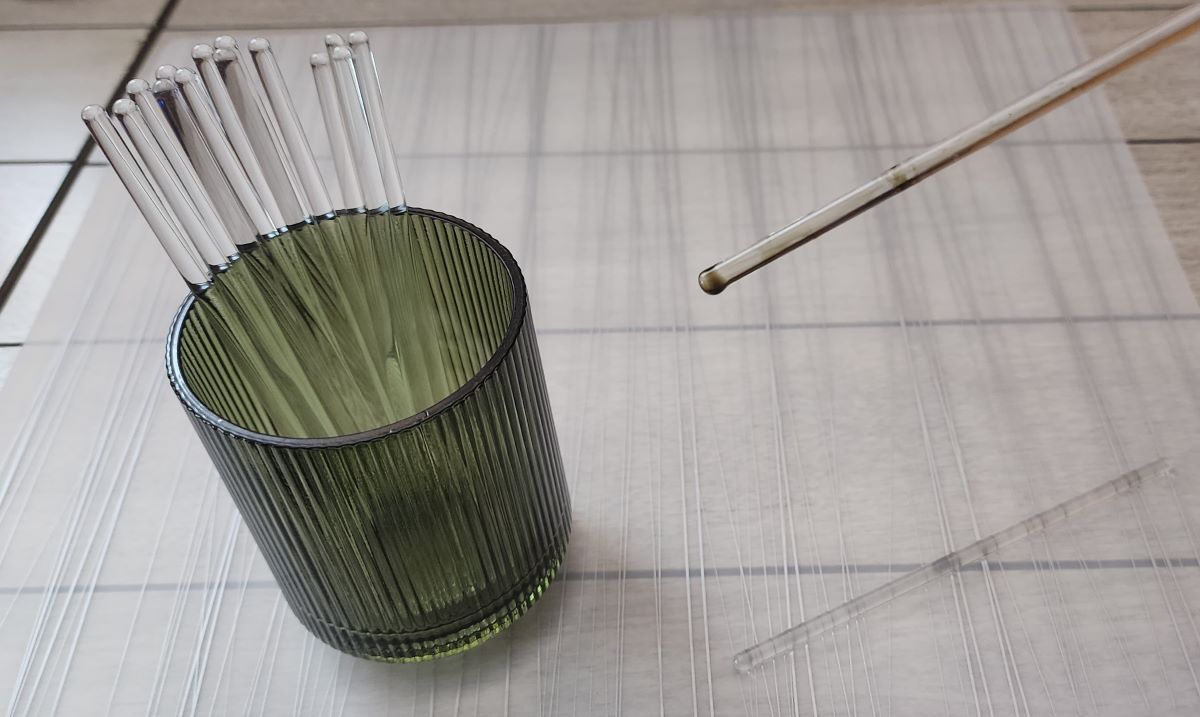 A green glass cup with a handful of thin glass stir rods with rounded ends, and one glass stir rod held in the air after being dipped in ink.