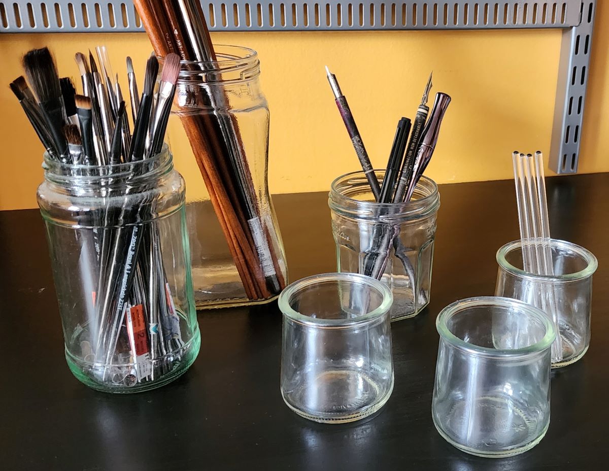 Several tall food jars full of paint brushes, dip pens, etc. and three smaller glass jars with a slightly tapered shape and plain lip at the top.