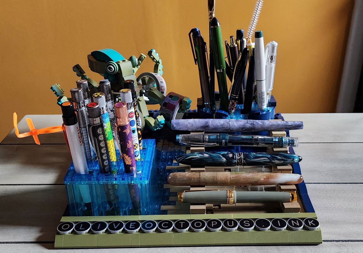 A custom LEGO desk tray with a dark blue base, olive green octopus in the back left corner holding rolls of washi tape on a few tentacles, a rounded and divided pen/pencil/marker/scissor storage area in the right back corner, a tall transparent blue grid of 9 pen slots for Retro 51 pens and pencil in the front left corner, and tiers of horizontal pen stands in tan bricks holding fountain pens in the front right corner. Round flat pieces with letters spell out oliveoctopus.ink across the front.