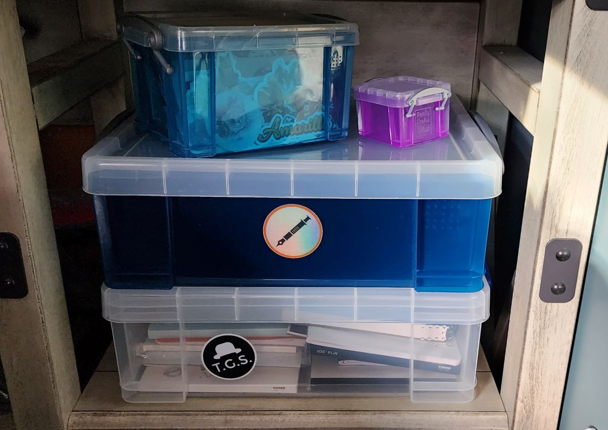 A stack of four plastic boxes with lids, one clear medium box filled with notebooks and paper, a second medium box in blue with a clear lid, a smaller blue box filled with stickers, and a mini purple box.