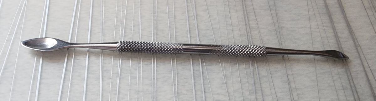 A double-ended metal hand tool with knurled grip, one end has a small, slightly pointed spoon shape with a thin edge on one side, and the other end is a narrower shape that ends in a sharper point.