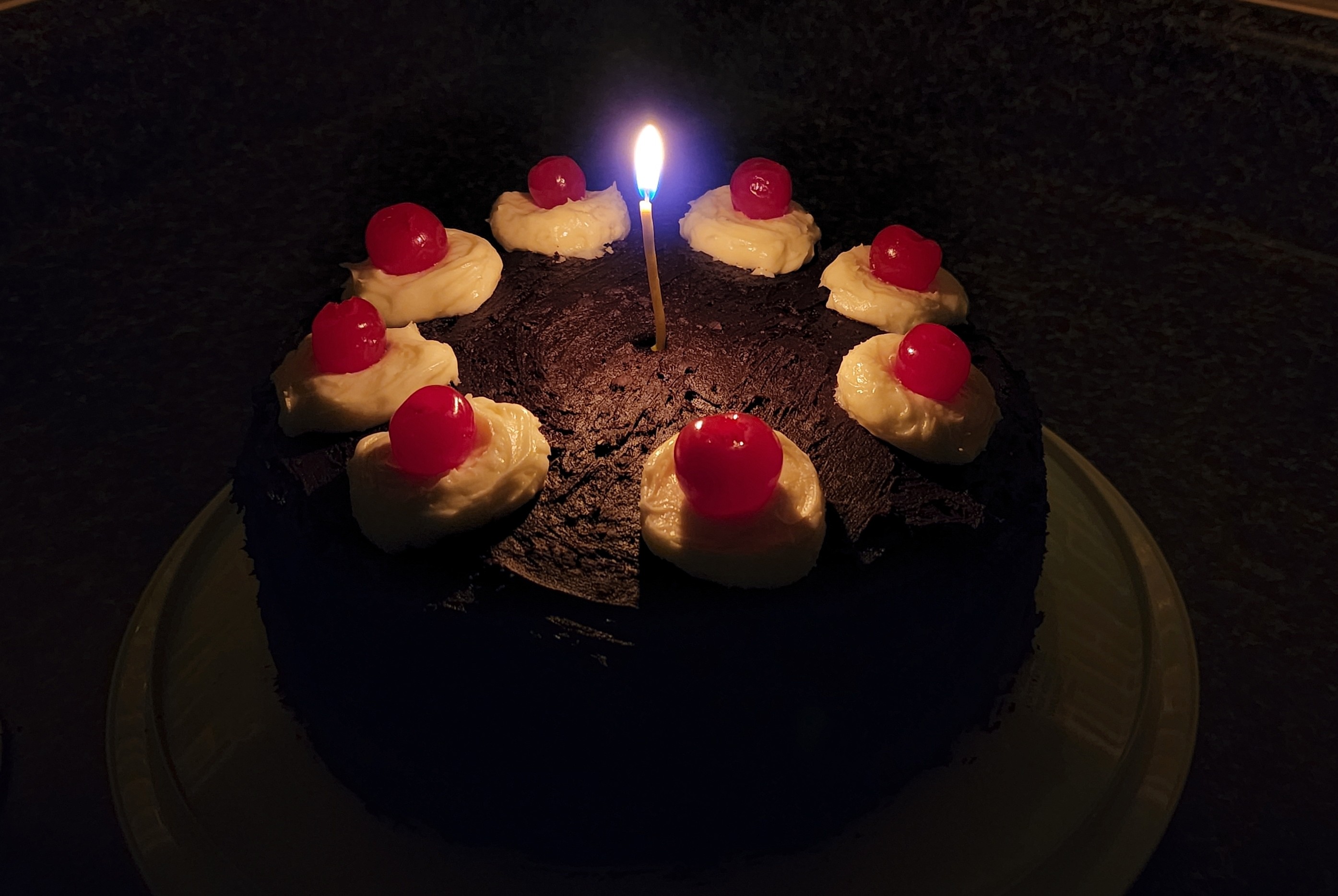 A round two-layer cake covered in chocolate frosting and decorated with a circle of cherries on circles of white frosting with a lit candle in the middle.