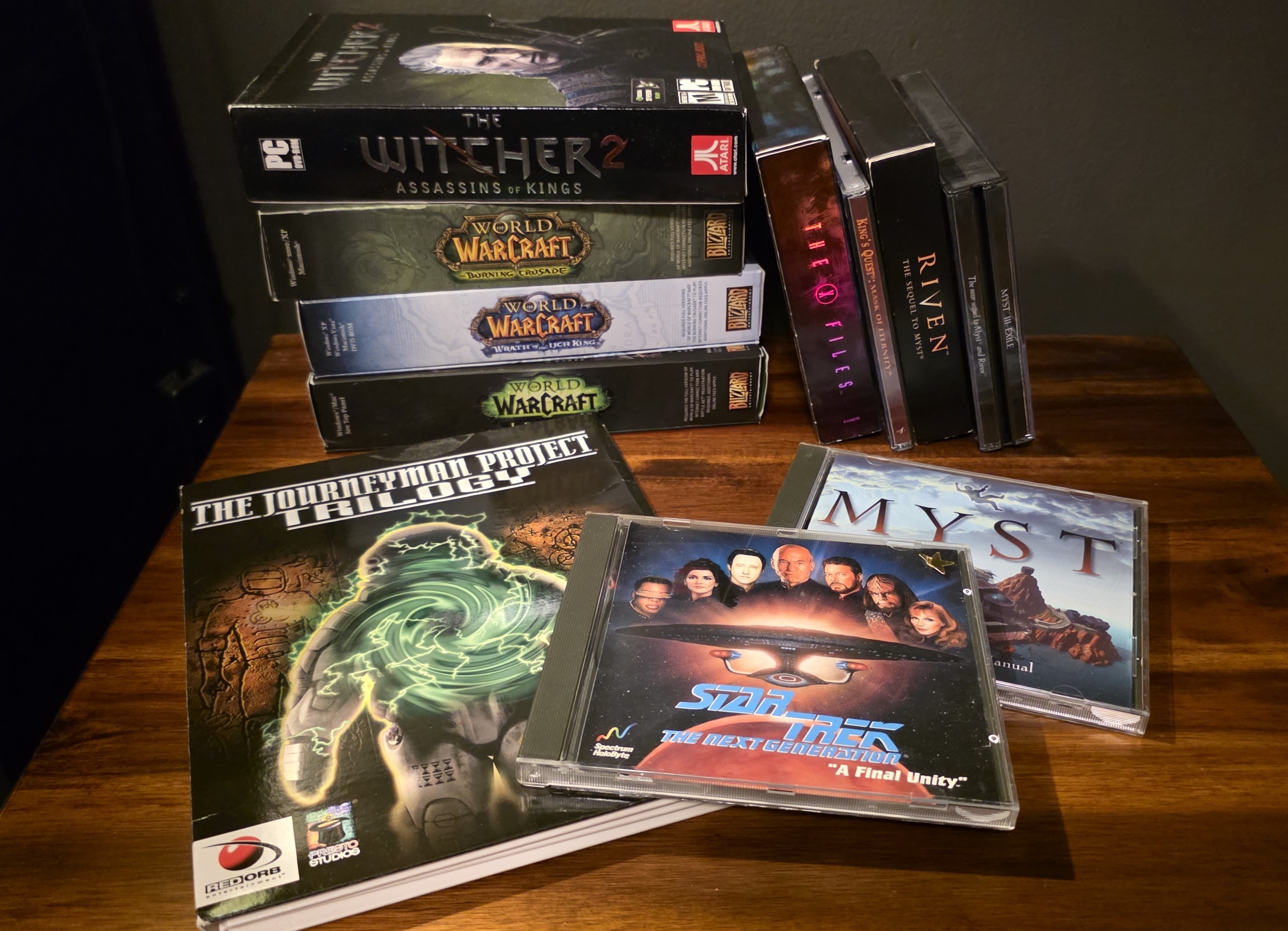 Boxes and jewel cases with discs for various old computer games featuring The Journeyman Project Trilogy, Star Trek The Next Generation 'A Final Unity', and Myst in the foreground, with Riven, Myst III, King's Quest, The X Files, Witcher 2, and three World of Warcraft expansions in the background.