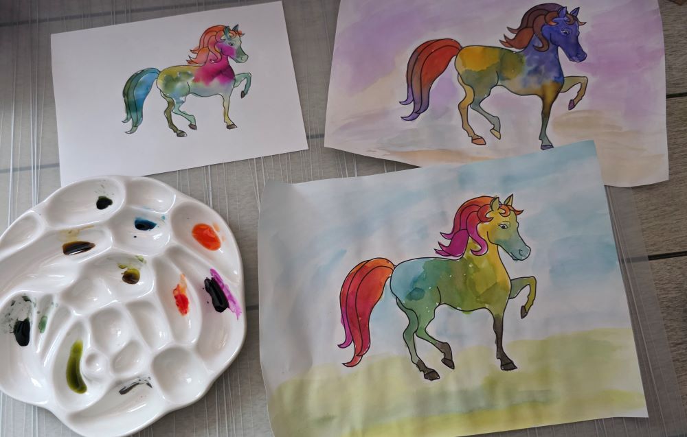 Coloring book outlines of ponies painted in with a rainbow of colors and a ceramic paint palette.